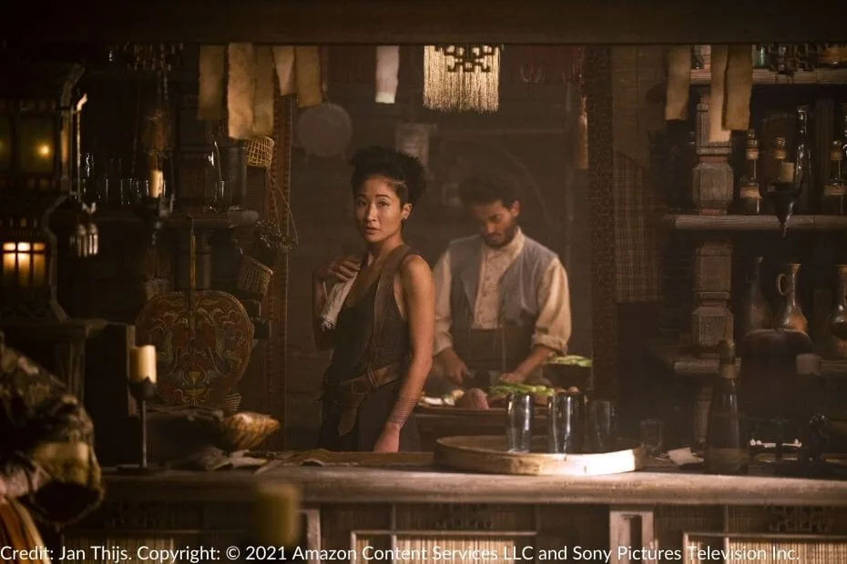 Min stands behind the bar in Fal Dara, her expression pensive as she gazes forward. The dimly lit room is filled with hanging lanterns, shelves of bottles, and an air of quiet intrigue.