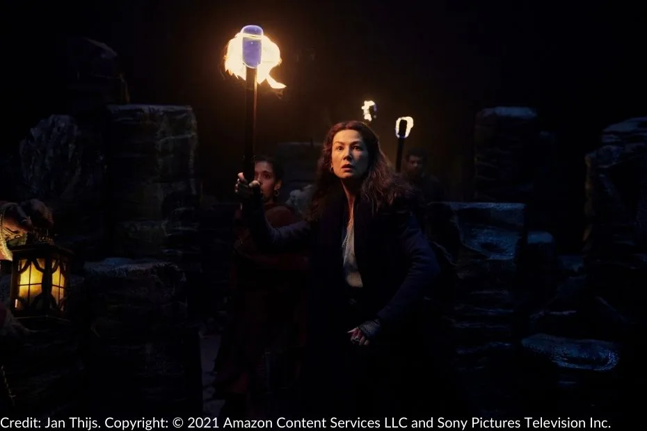 Moiraine holds a flaming torch in the dark, surrounded by jagged stone formations of the Ways. Her expression is intense as she looks ahead, with other figures visible in the shadows behind her.