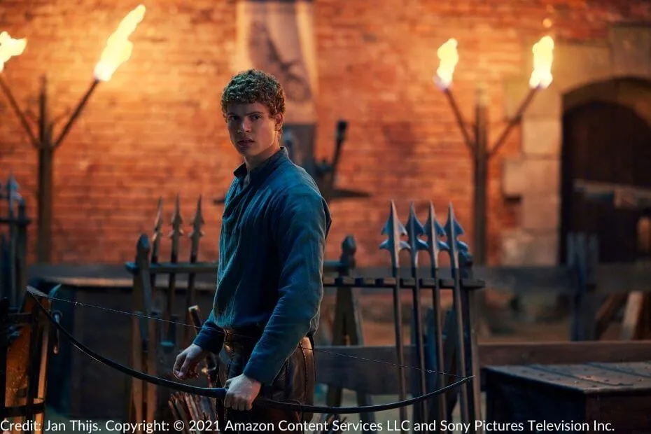 Rand stands in a torch-lit courtyard, holding a bow by his side. His intense gaze is fixed forward, ready to take aim. Spears are lined up behind him.