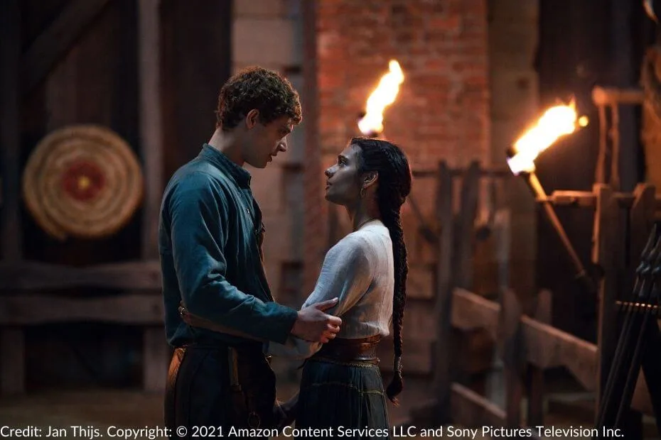 Rand and Egwene stand close in a torch-lit courtyard. Rand holds Egwene’s arm while she rests her hand on his waist. A target and weapon racks are visible in the background.