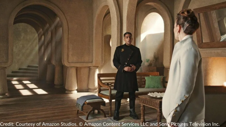 Lews Therin stands in a sunlit room, wearing a black coat with a gold dragon pin, passionately pleading with Latra Posae, who listens intently in a white suit.