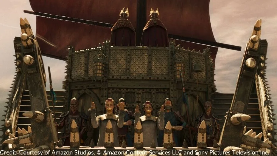 A massive Seanchan ship with ornate details approaches. Two women, their mouths covered by metallic gags, raise their hands in a gesture, while others stand stoically behind them, flanked by armored soldiers with upright pikes.