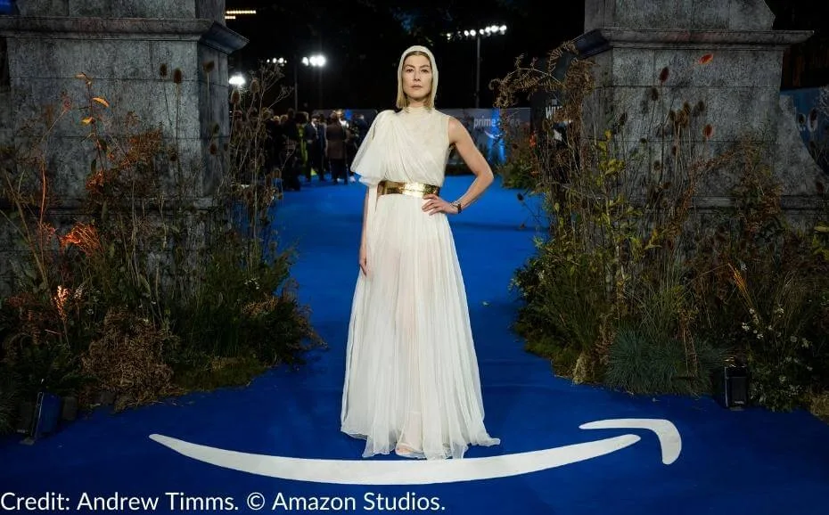 Rosamund Pike, dressed in an elegant white gown with a golden belt, attends The Wheel of Time Season 1 premiere, standing on a blue carpet surrounded by nature-inspired décor and the base of the show's Waygate.