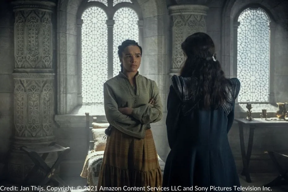 Nynaeve stands with arms folded, looking unimpressed as Moiraine Damodred speaks to her in a sunlit chamber of the White Tower.