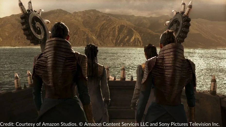 Four Seanchan figures stand on the deck of a ship, facing the mountainous coastline. Their elaborate armor, sharp ridges, and towering metallic emblems mark them as part of an invasion. The ocean stretches before them.