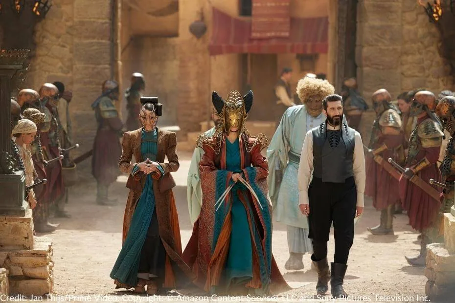 Lady Suroth and Alwhin walk through Falme, dressed in regal Seanchan attire, their faces partially concealed by elaborate masks. Ishamael strides beside them while Loial follows closely behind. Rows of Seanchan soldiers stand at attention, their armor gleaming under the sun.