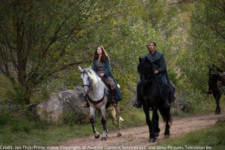 Moiraine and Lan ride side by side along a forested path, their expressions solemn. Moiraine sits atop a white horse, her blue cloak draped over her shoulders, while Lan rides a black steed, ever watchful.